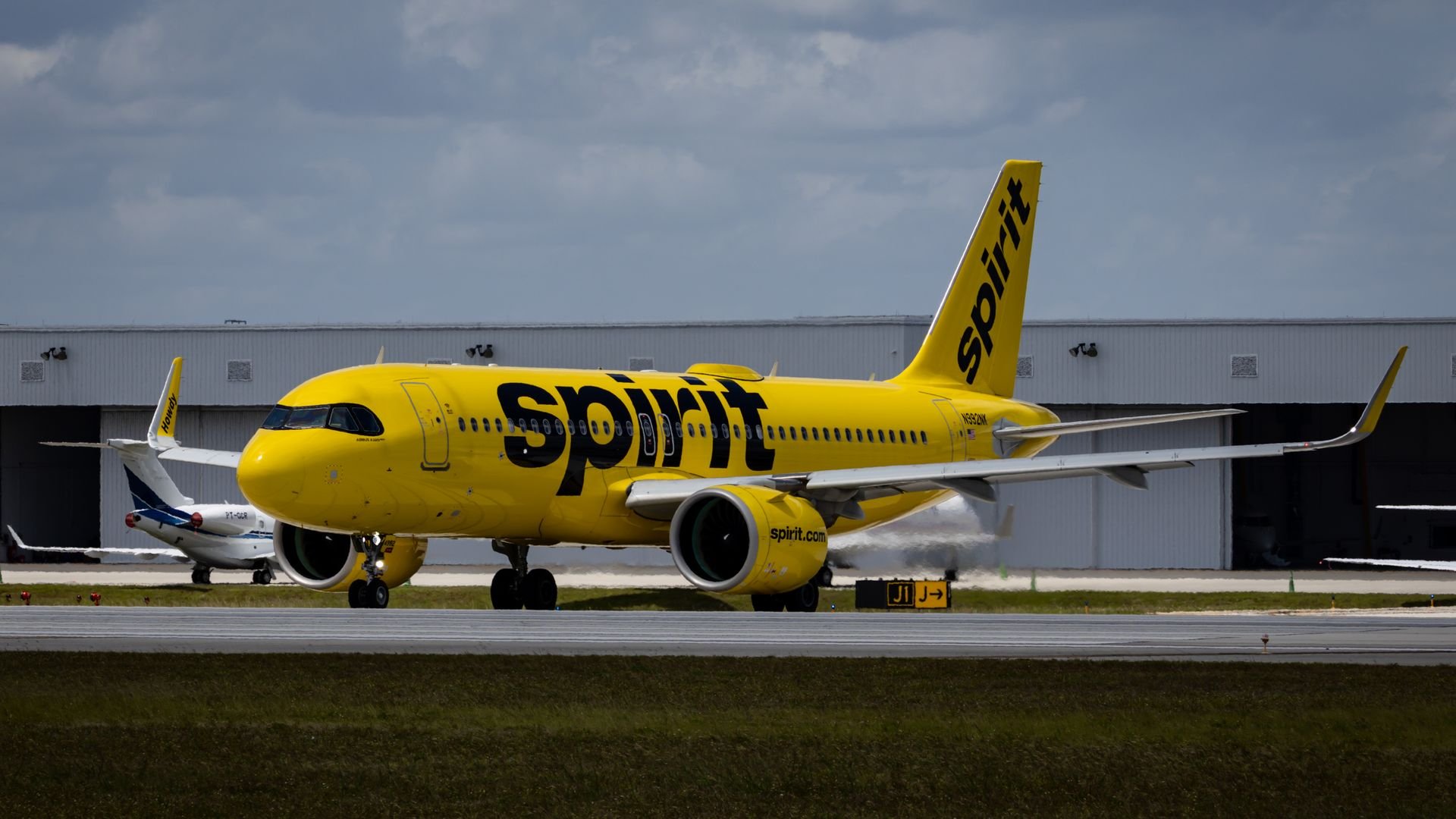 A Spirit Airlines plane, yellow with the word SPIRIT painted on the side, sits in front of an airport hangar. 