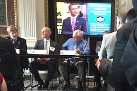 Gregory Olson and other panelists sit at table under a monitor featuring Barack Obama at the White House.
