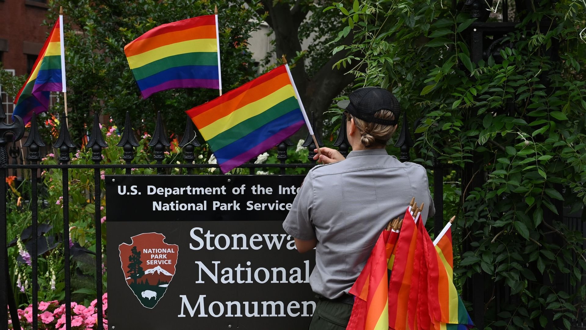 A person wearing a gray shirt and army green pants puts rainbow flags along a fence adorned with a sign that reads 