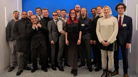 Stop Killing Games A large group of people standing and smiling in suits for a photo in front of a blue background, including Ross Scott, founder of Stop Killing Games