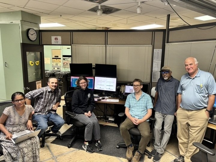 Six people pose with a computer and several monitors in a small office