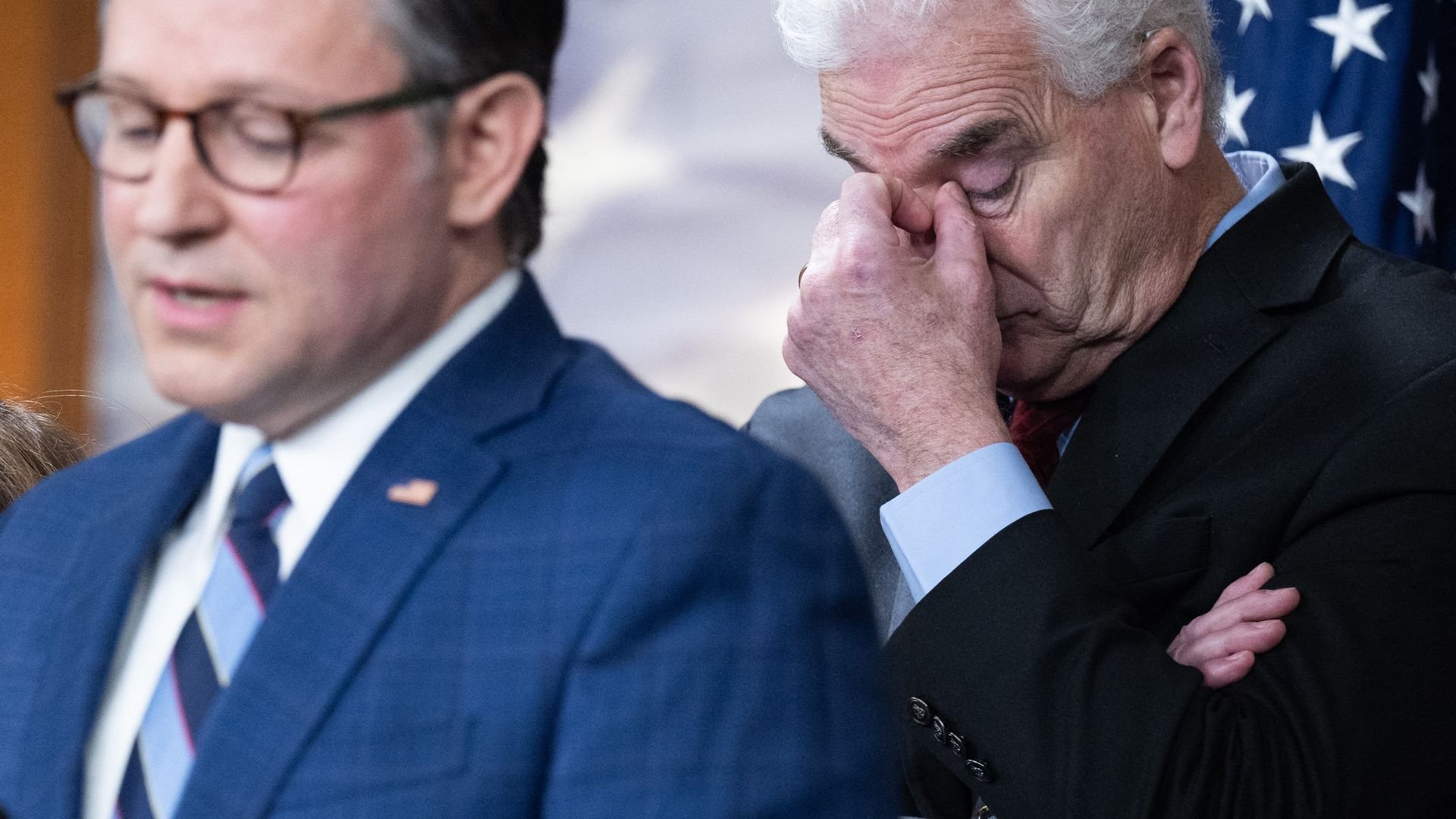 Two men in suits at a formal event; one in a blue checkered suit speaking, the other in a black suit with white hair rubbing his eyes, with an American flag in the background.
