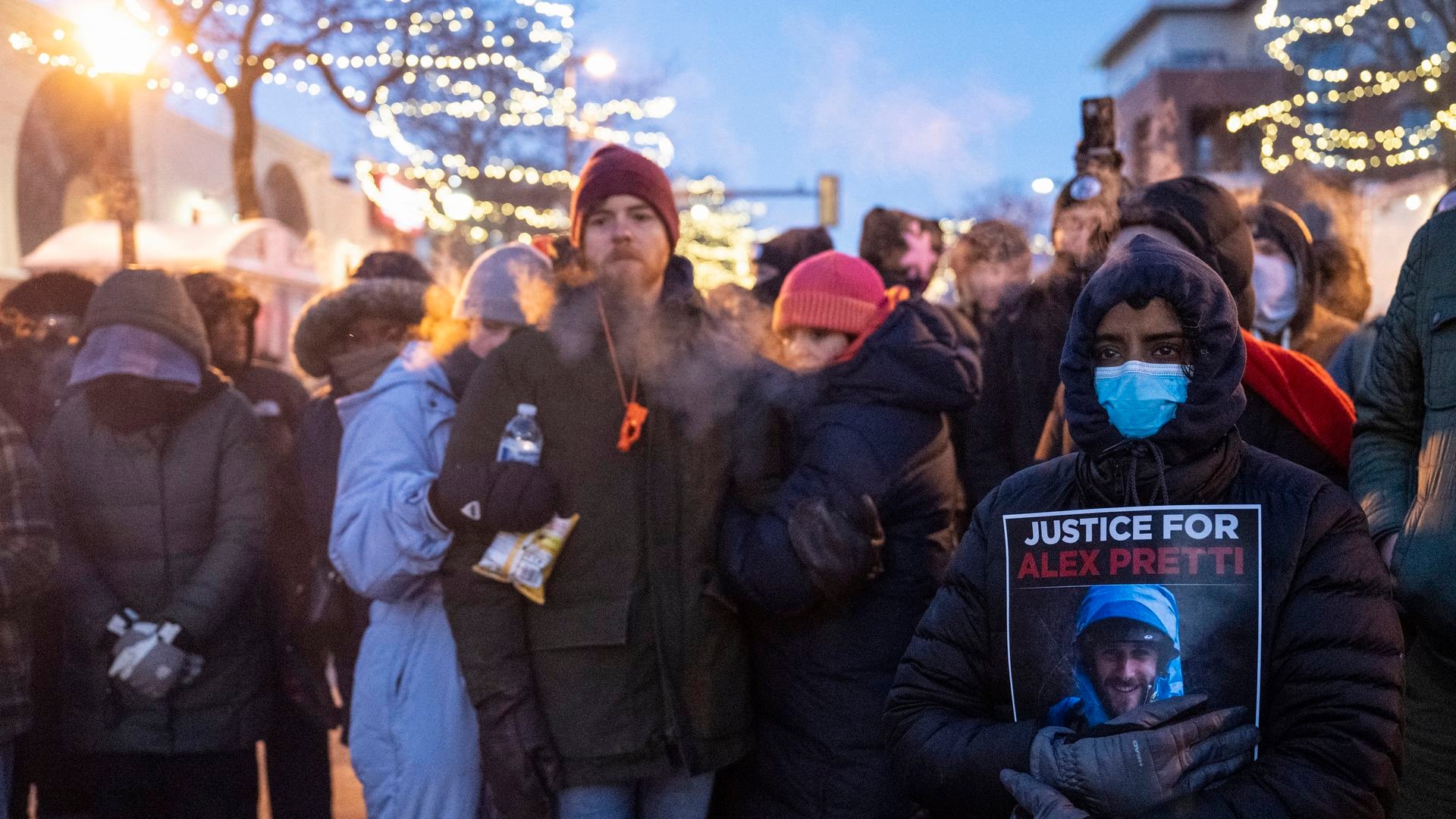 A group of people stand together at a makeshift memorial for a man shot by federal agents in Minnesota. One person, wearing a blue mask, holds a sign that says