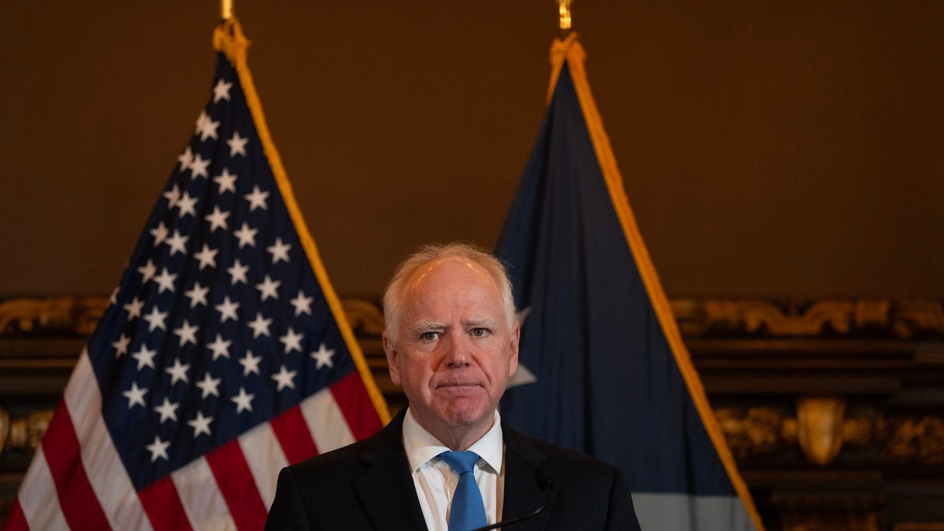 Walz standing at a microphone with a straight expression wearing a navy suit jacket, white collared shirt and light blue tie 