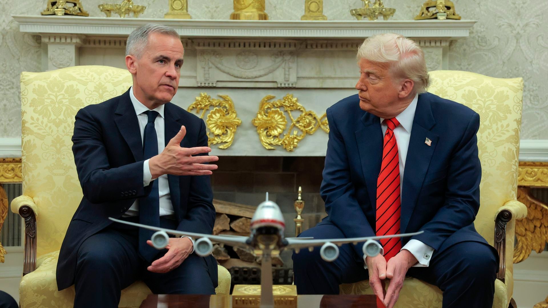 Mark Carney (left) and Donald Trump (right) seated in chairs at the White House with a model airplane between them and ornate background