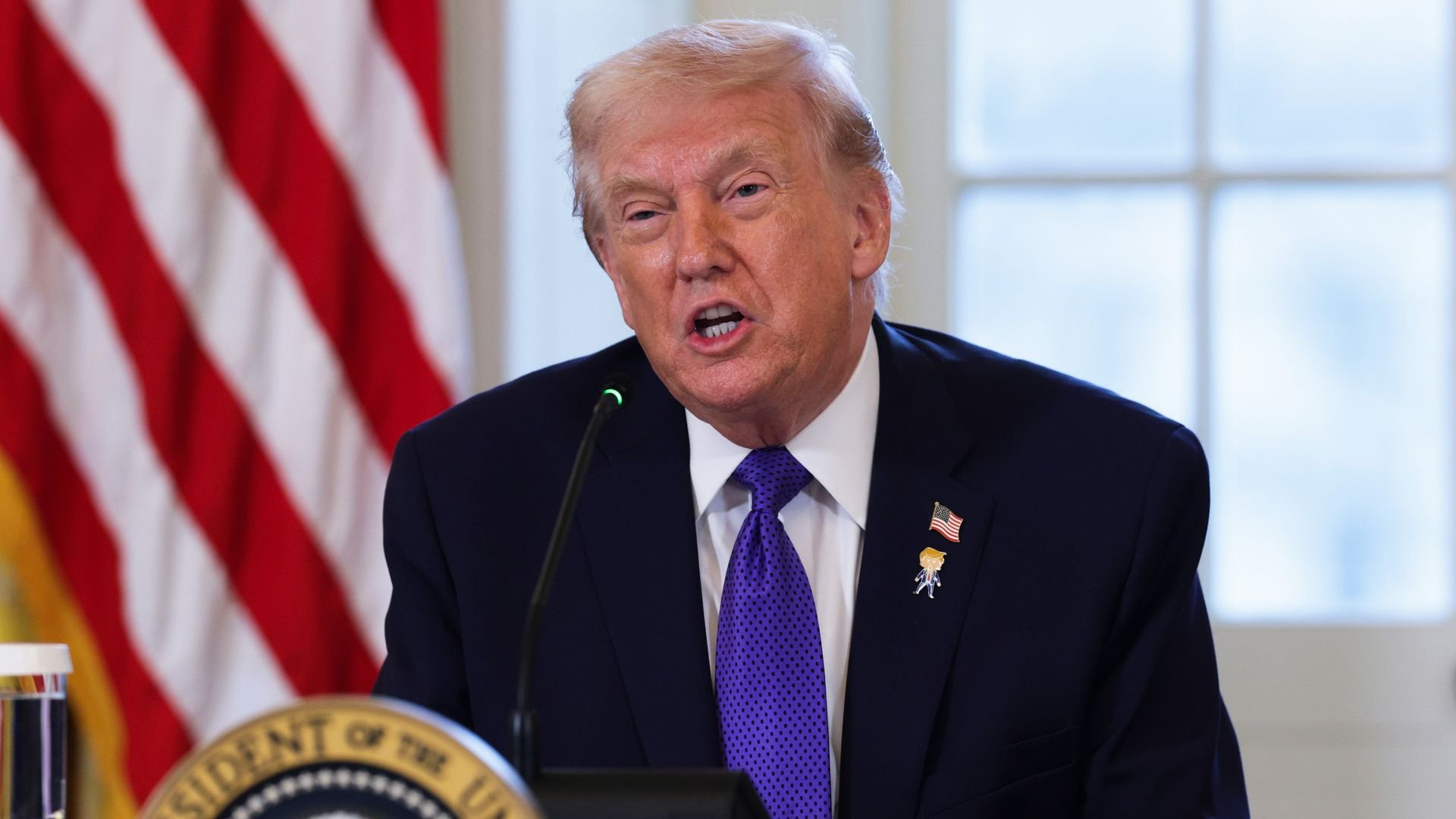 President Trump, with light hair wearing a navy suit and purple tie speaks into a microphone, with a blurred U.S. flag and a presidential seal visible in the background.