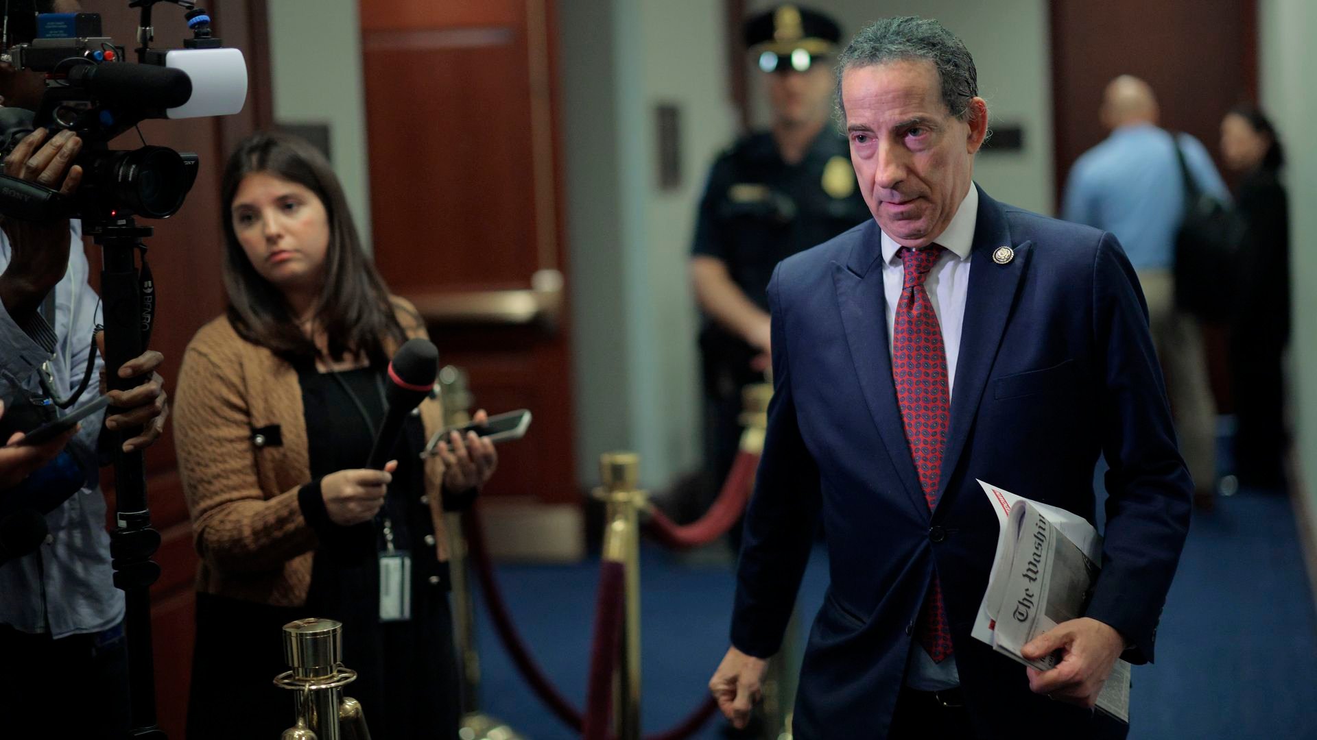 Man in a dark blue suit and red tie holding a newspaper walking past a reporter with a microphone and camera in a hallway with blue carpet and a police officer in the background.