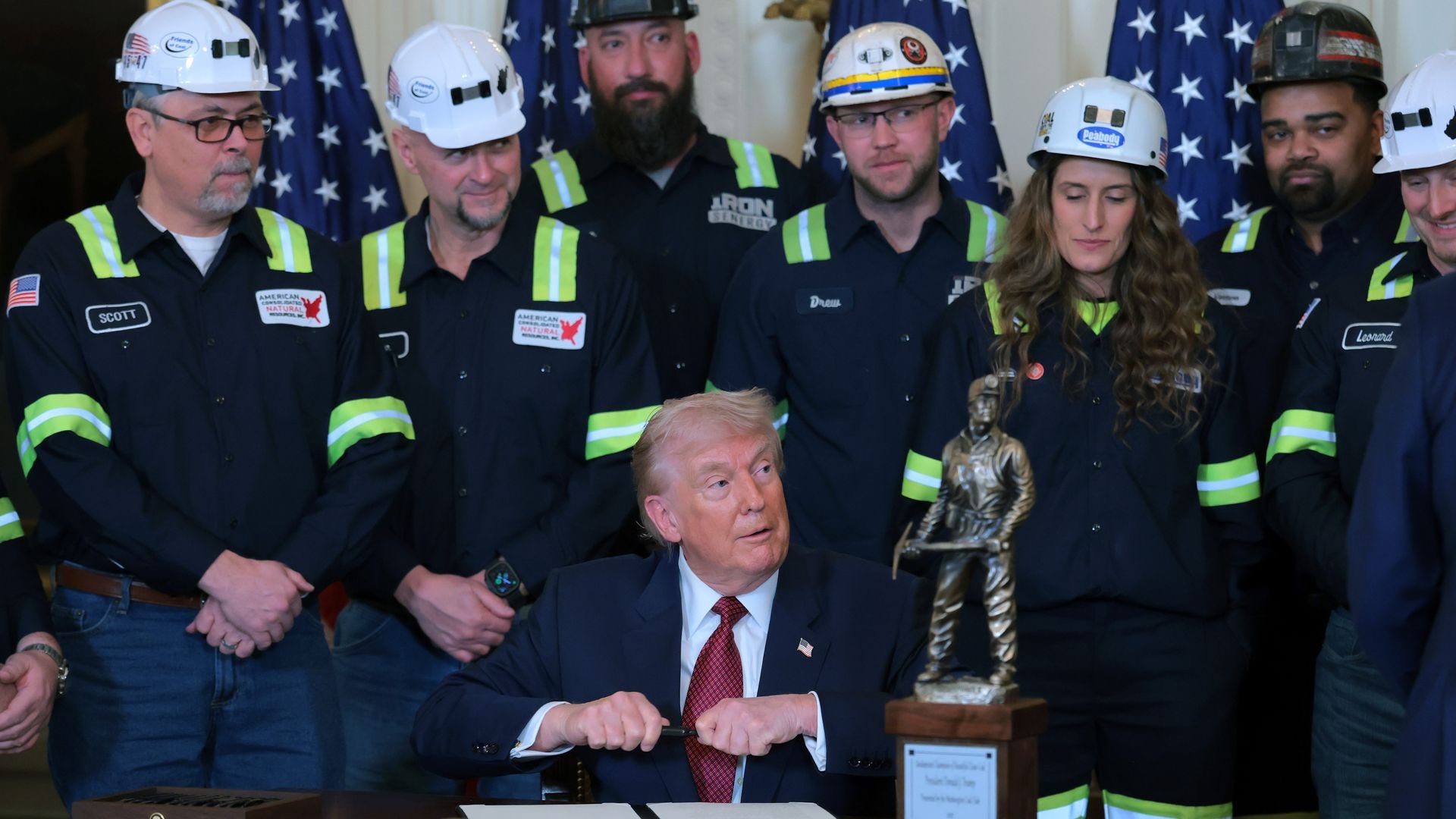 President Trump in a dark suit signing a document, surrounded by workers in dark uniforms and white helmets, with American flags in the background and a miner statue on the desk.