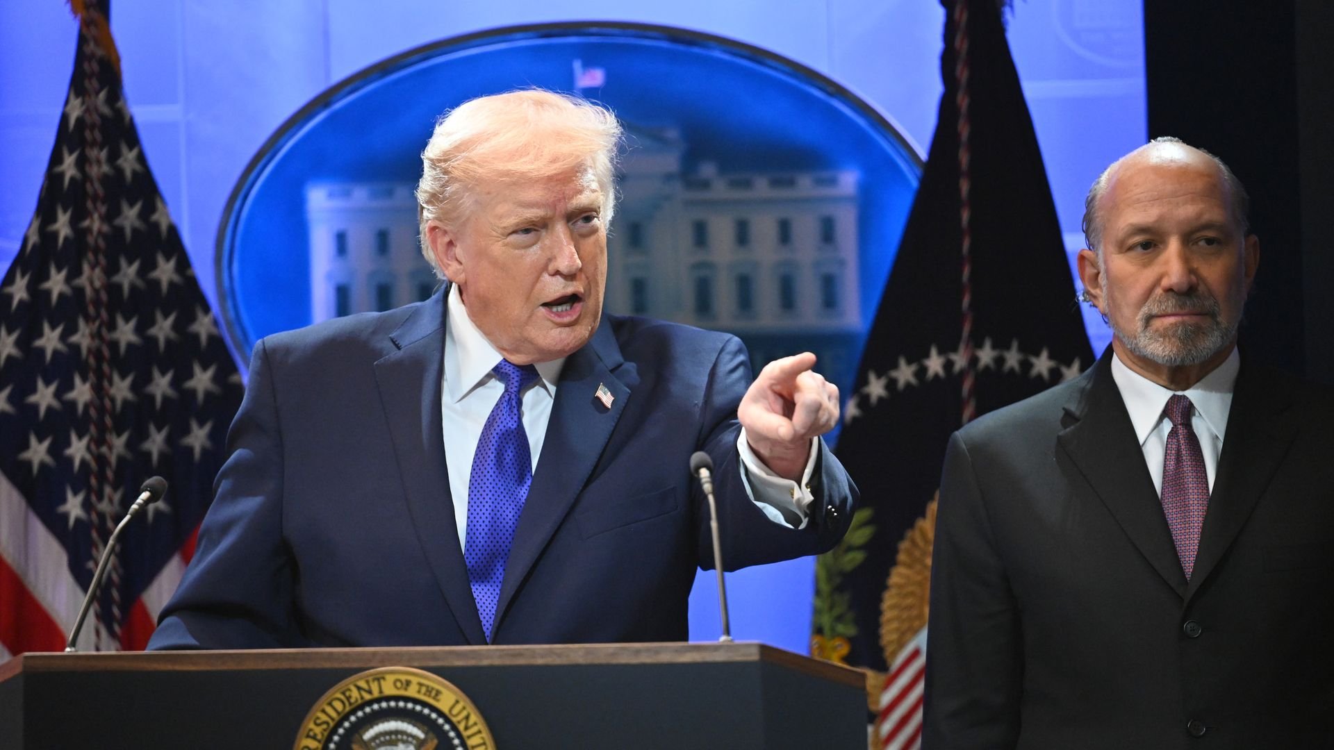 Man points while at podium and another stands to his side