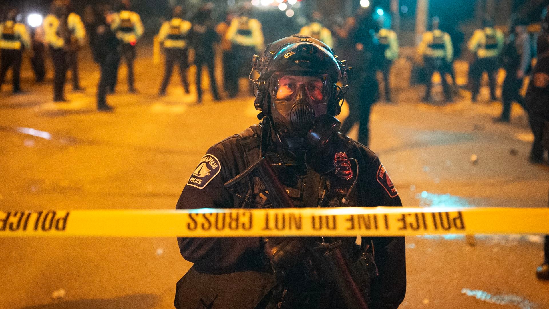 A police officer in riot gear wearing a gas mask stands behind yellow police tape with other law enforcement in the background at night on a city street.