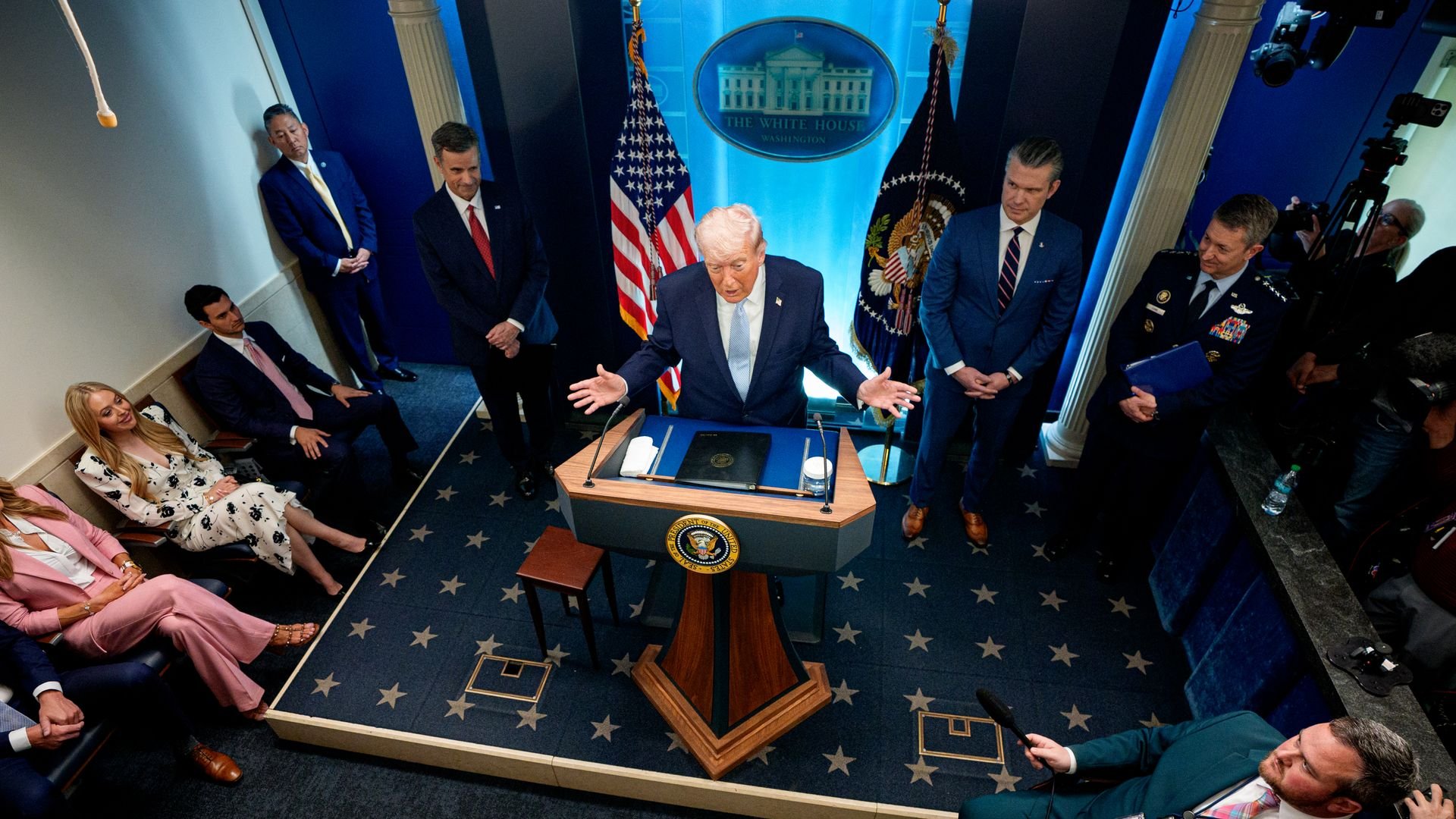 President Donald Trump speaks at a podium in the White House briefing room, surrounded by officials and reporters.