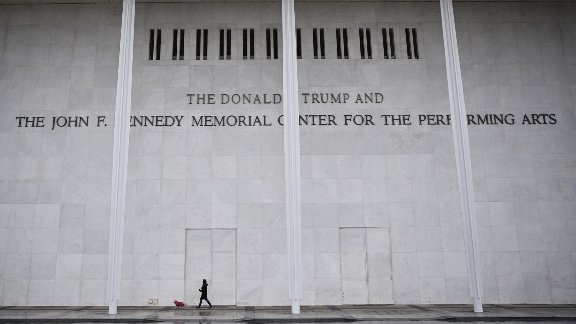 Woman walking a small dog in front of a large white marble wall with the inscription 