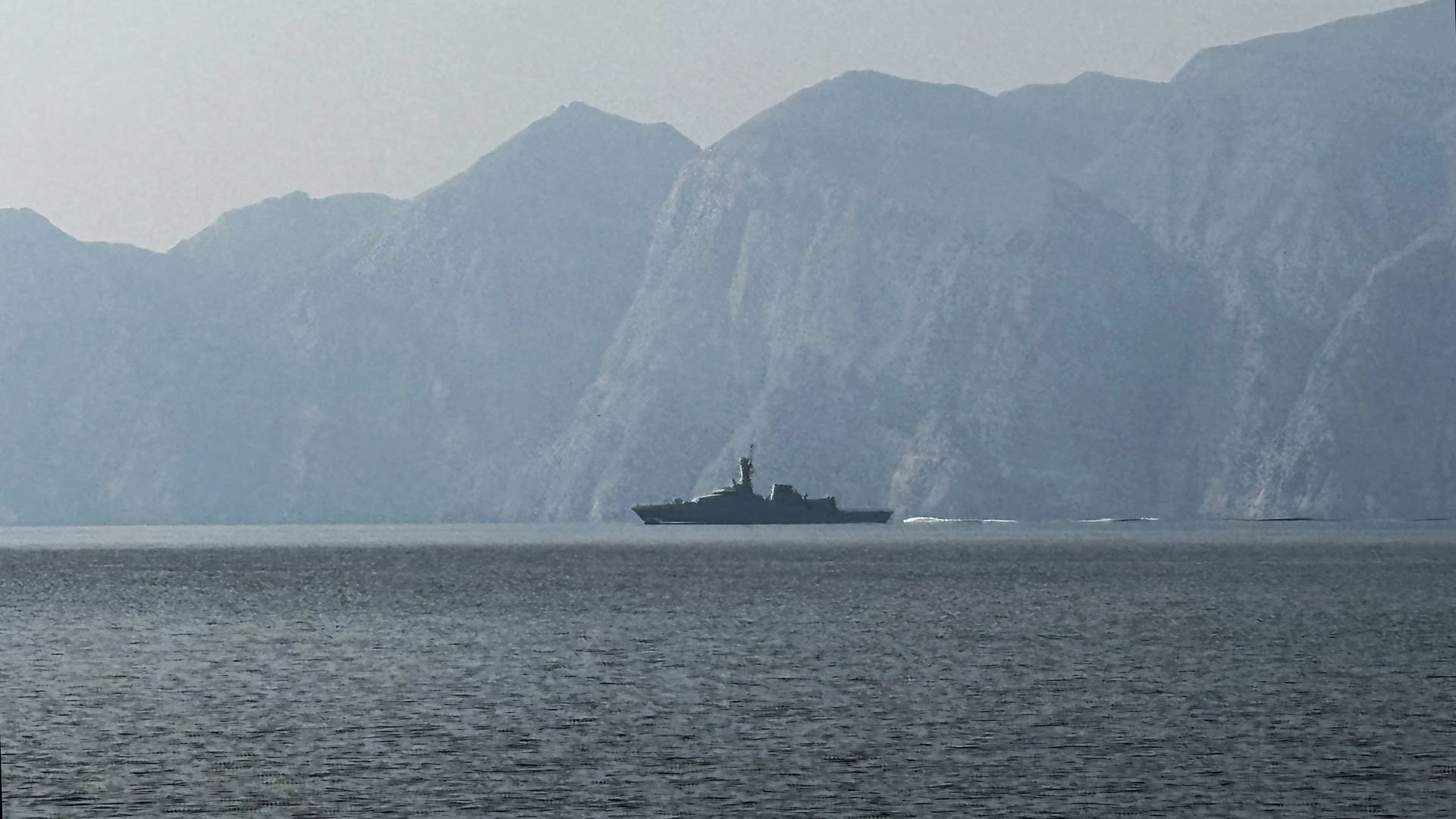 A military ship moves through the Strait of Hormuz with open water and a hazy sky in the background.