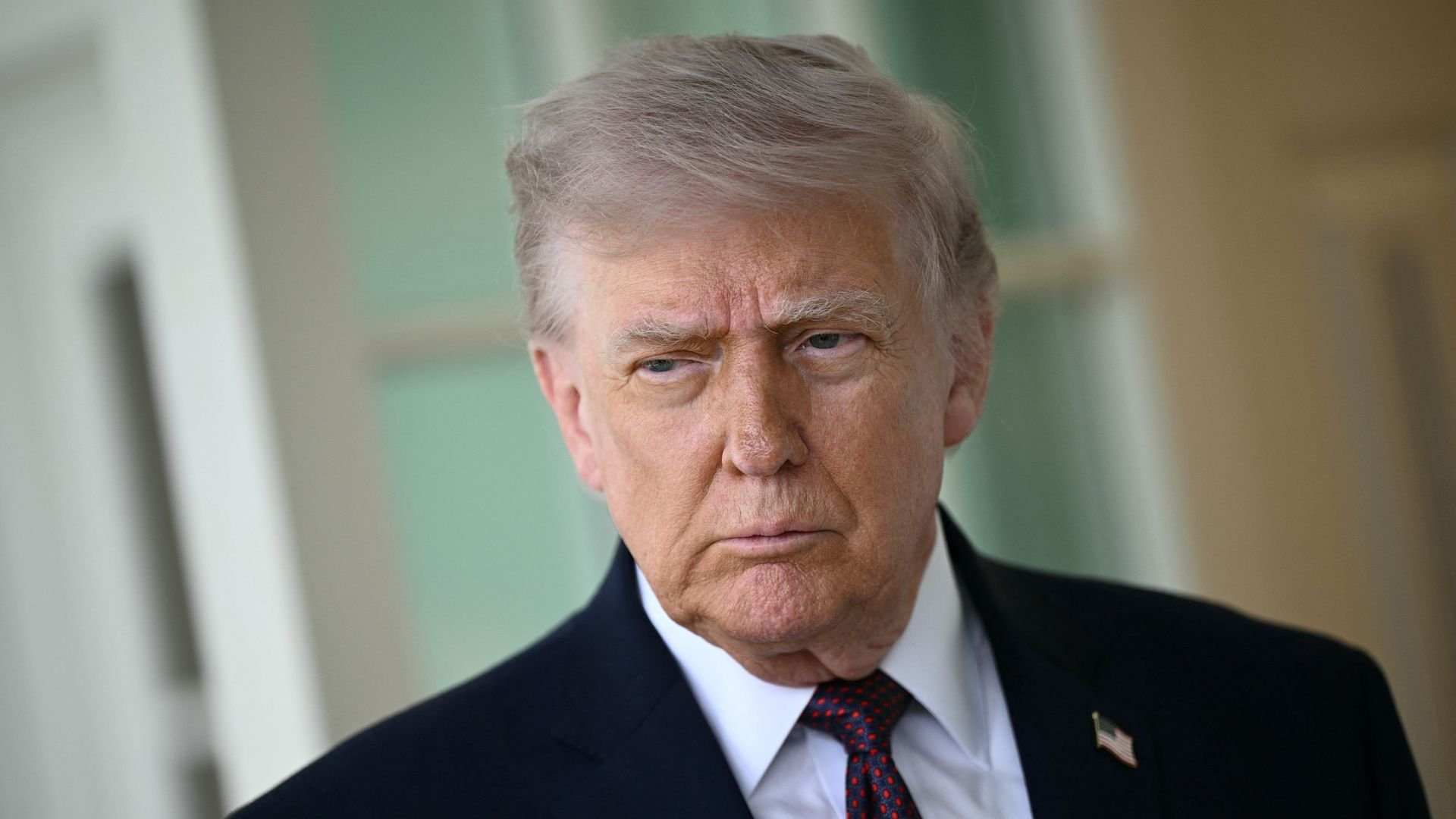 Close-up of President Trump  with gray hair in a dark suit, white shirt, and red tie, wearing a small American flag pin; he has a serious, furrowed expression.