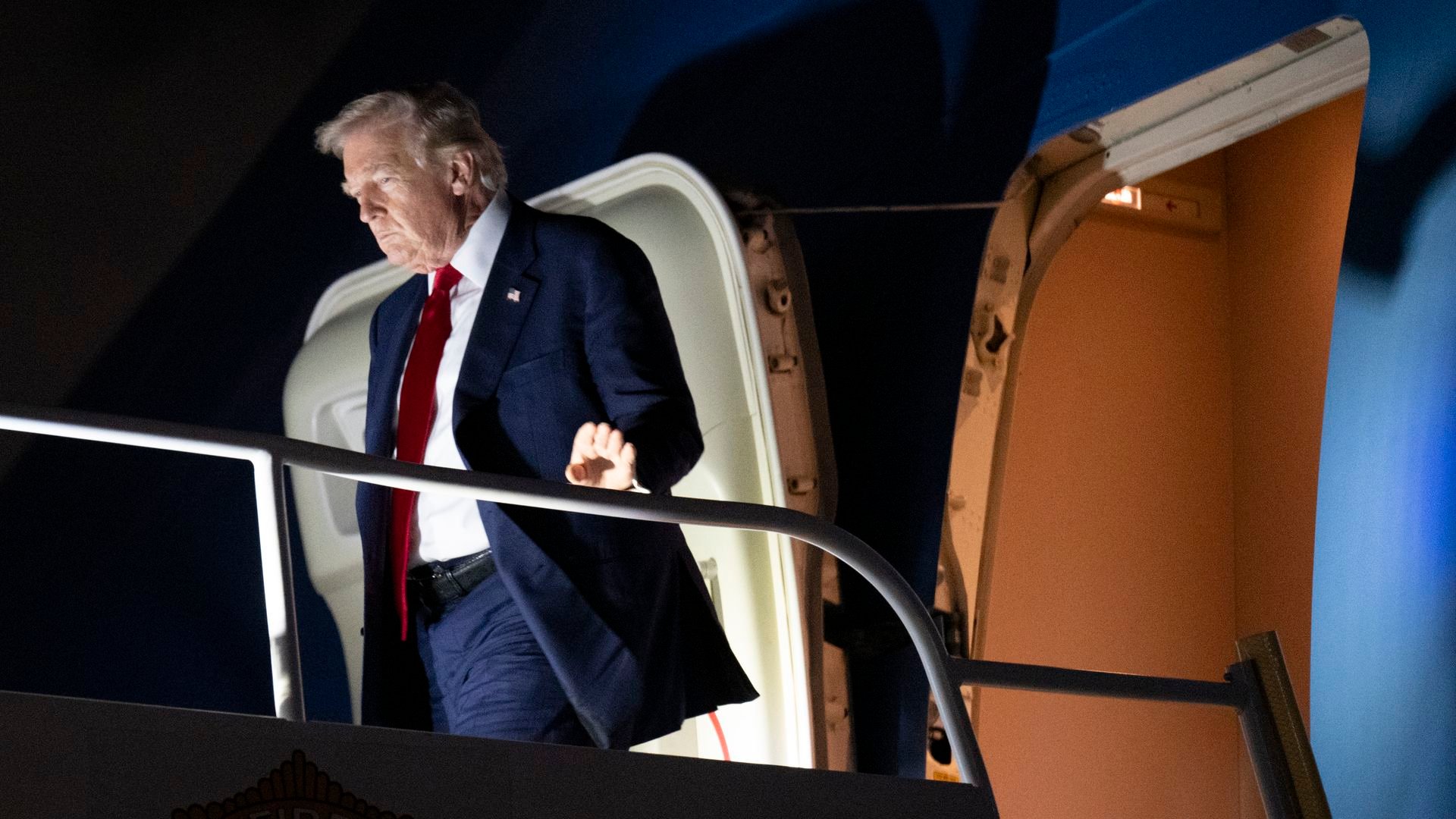 President Trump, illuminated by footlights against a dark background, exits an airplane. 