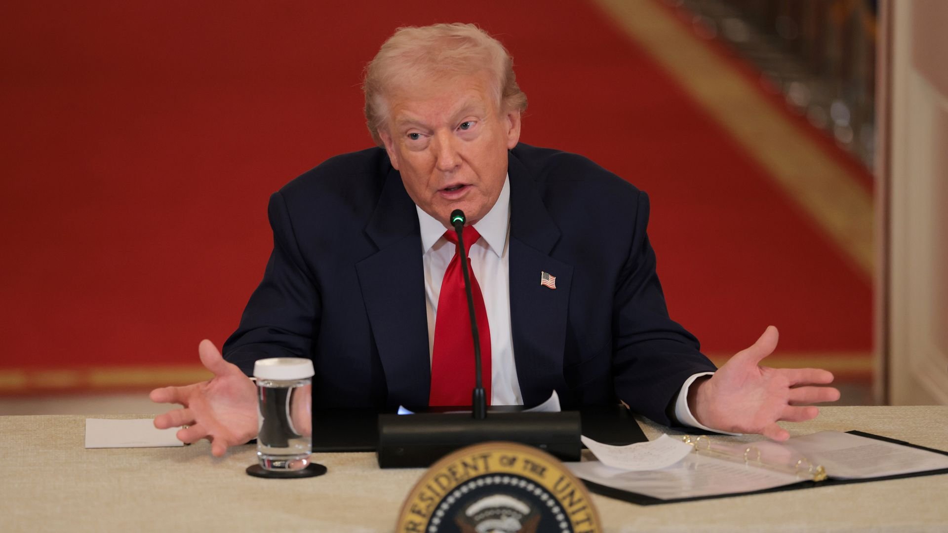 President Trump — wearing a dark suit jacket, an American flag pin, a red tie and a white collared shirt — holds his hands out as he speaks into a microphone during a roundtable discussion.
