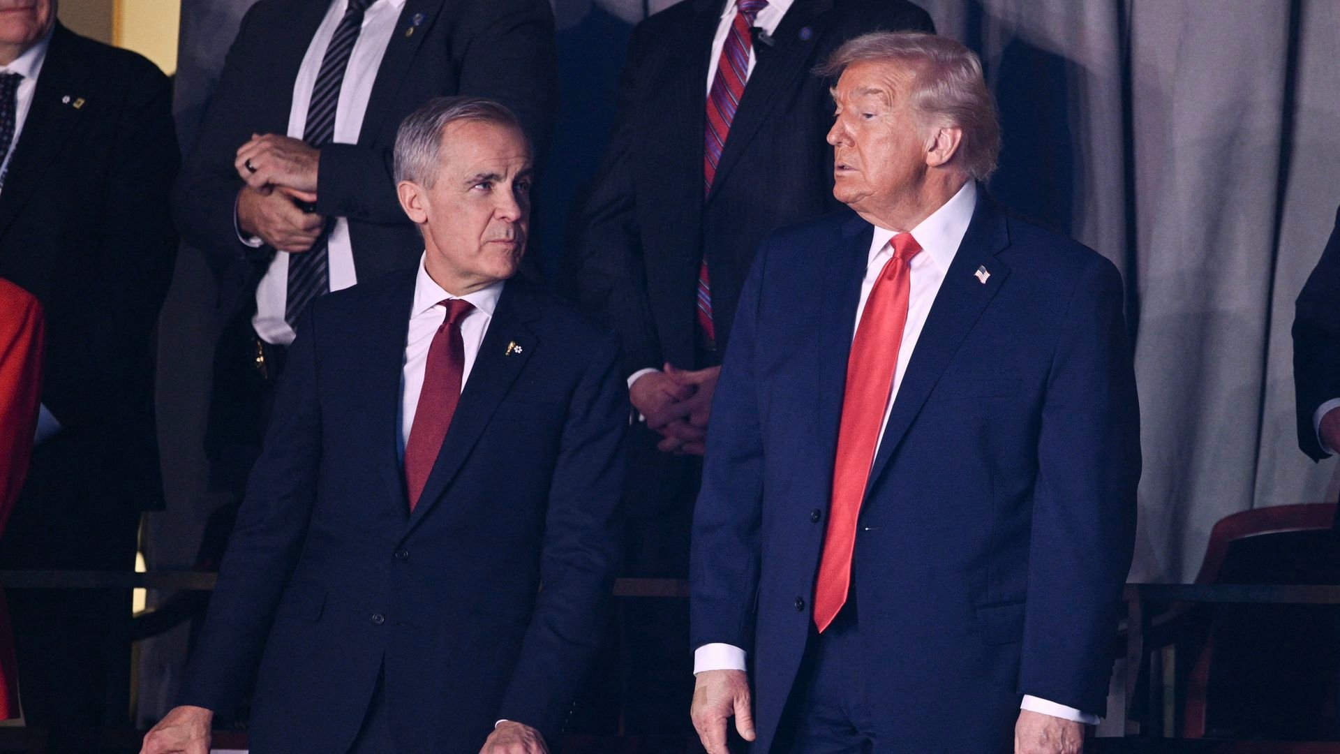 Two men in dark suits with red ties stand behind a maroon barrier. The man on the right has an American flag pin on his lapel. Other people in suits stand in the background.