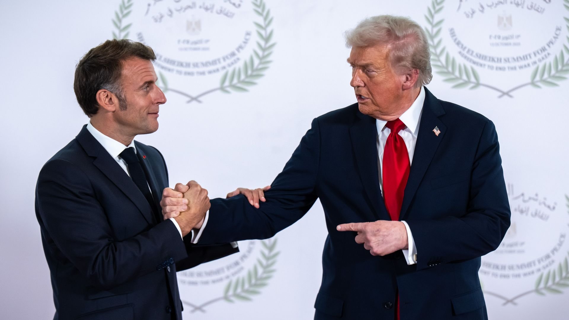 President Trump — wearing a dark suit, a white collared shirt, a red tie and an American flag pin — points at President Macron — wearing a dark suit, a black tie and a white collared shirt — as the two men shake hands.