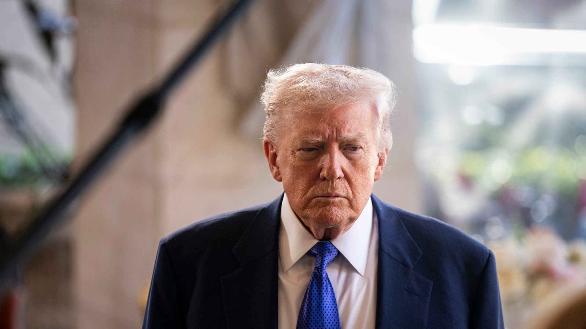 Trump with a straight expression wearing a navy suit jacket, white collared shirt and bright blue tie