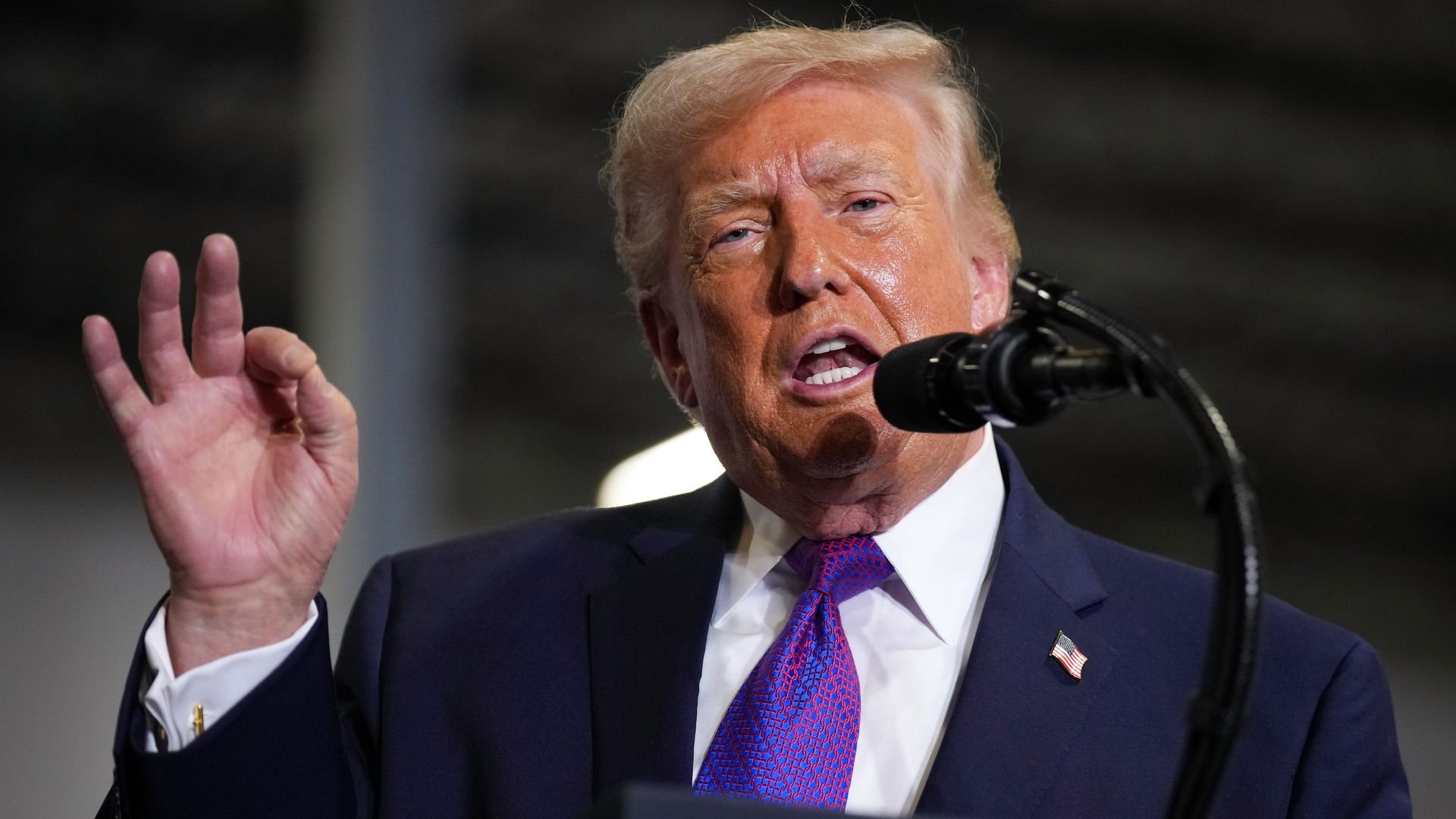 President Trump in a dark suit and white shirt speaks at a podium, wearing a purple patterned tie and an American flag lapel pin, gesturing with his left hand near a microphone.