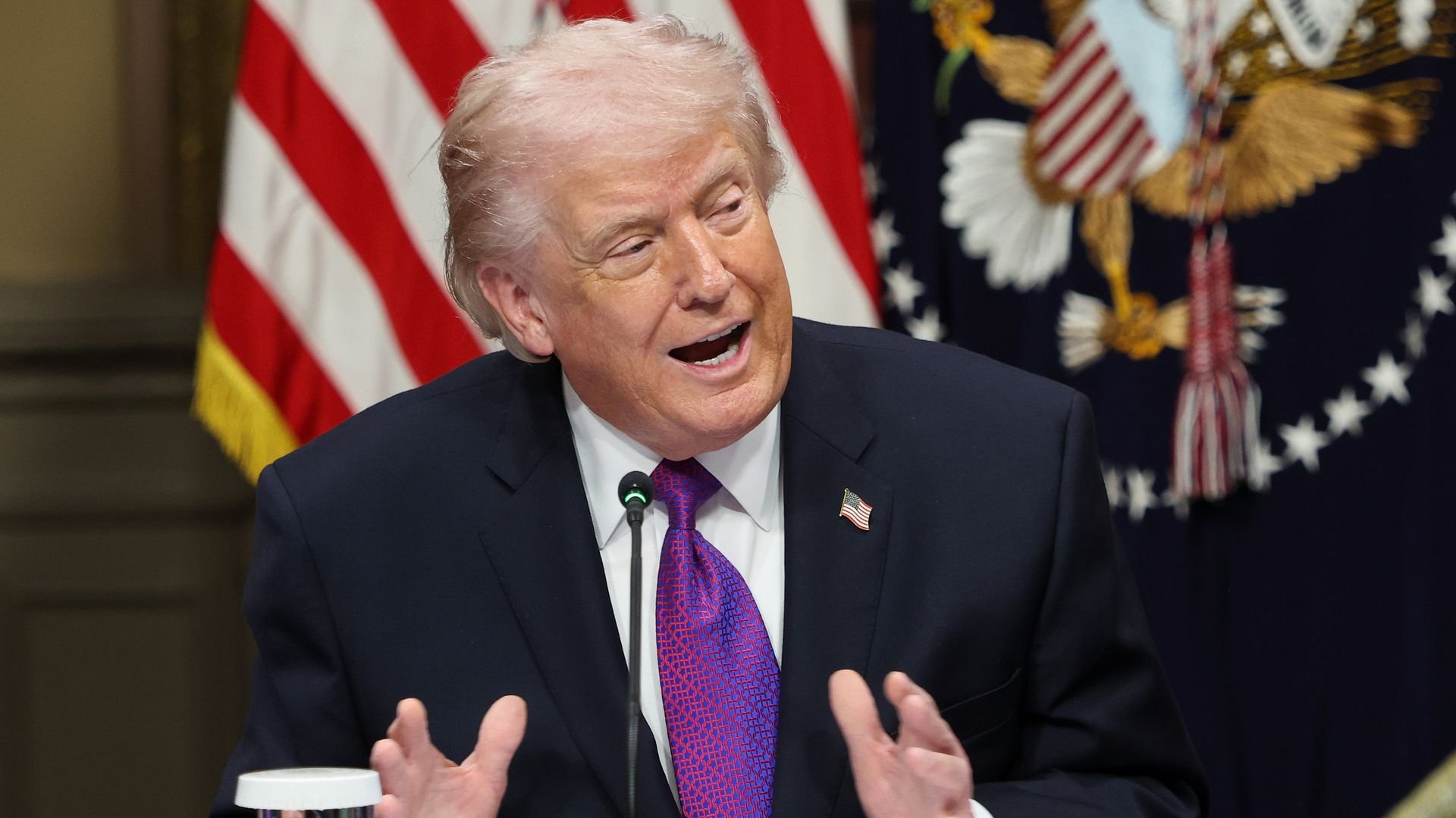 Trump speaking into a microphone with both of his hands raised to chest level, wearing a navy suit jacket with an American flag pin, white collared shirt and purple and blue design tie