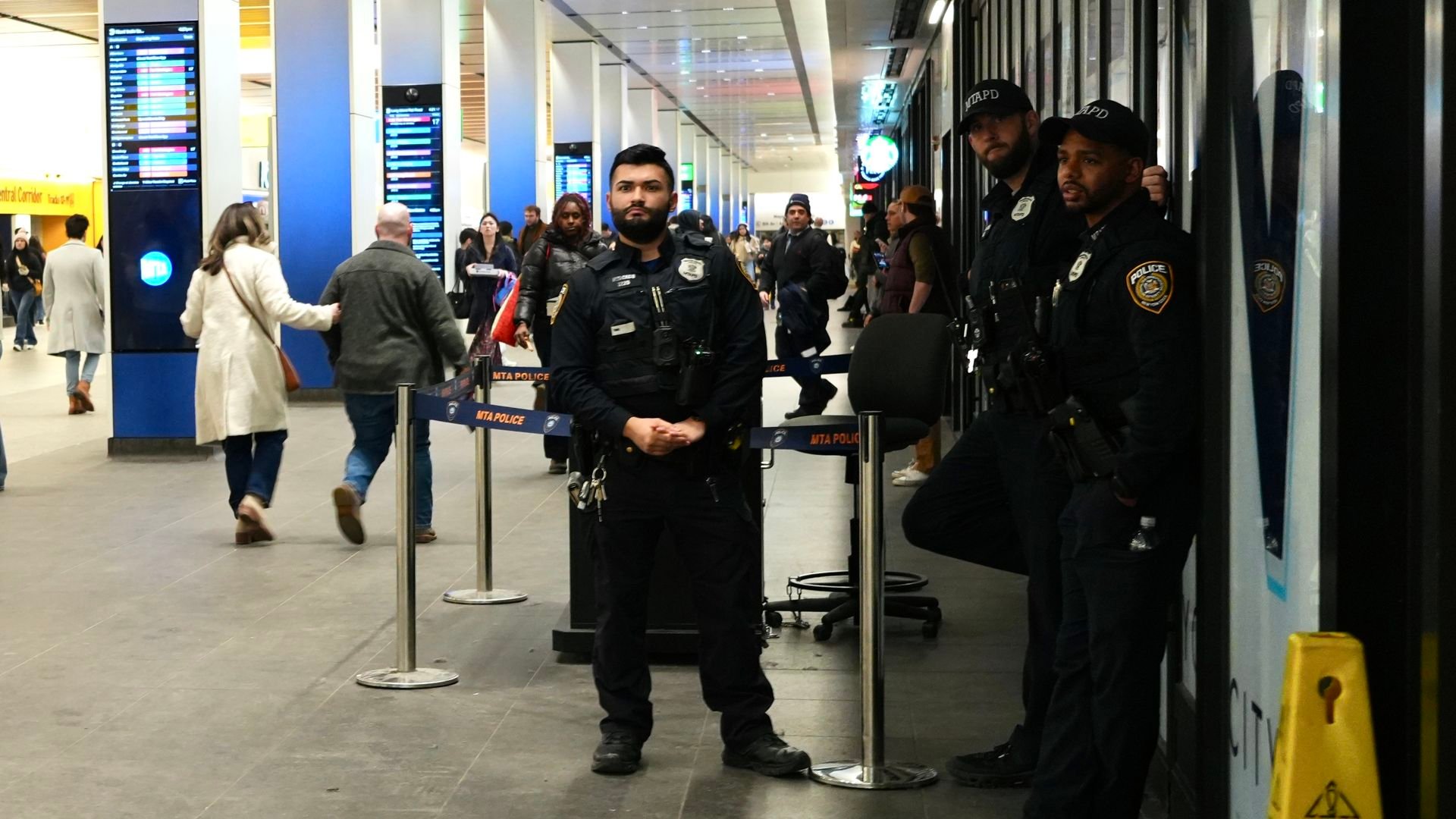 Three MTA police officers in uniform standing and leaning in a busy train station corridor with commuters walking by and digital schedule boards in the background.