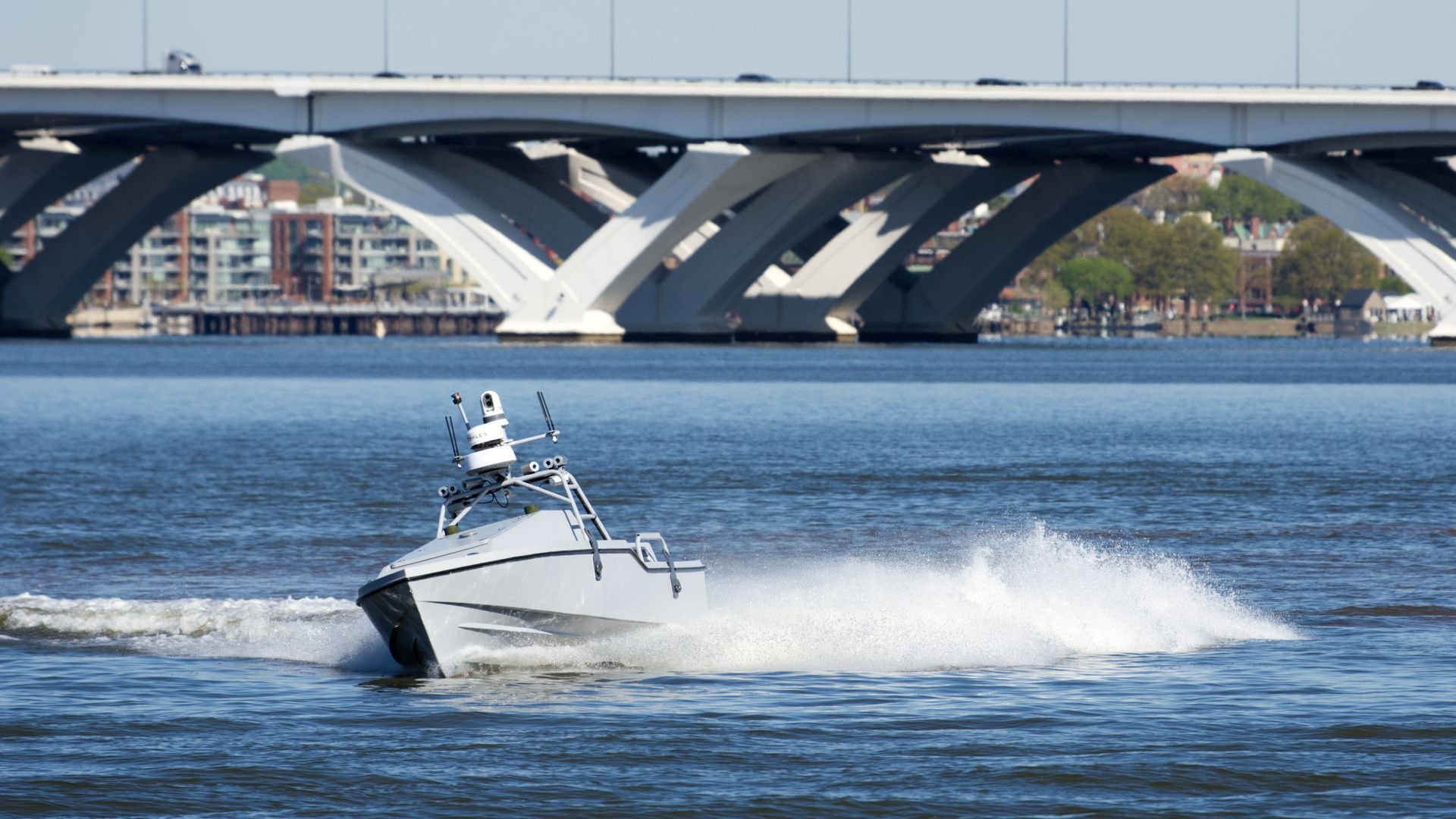 An unmanned boat cuts across the Potomac River. It's kicking up wake. A bridge can be seen in the background.