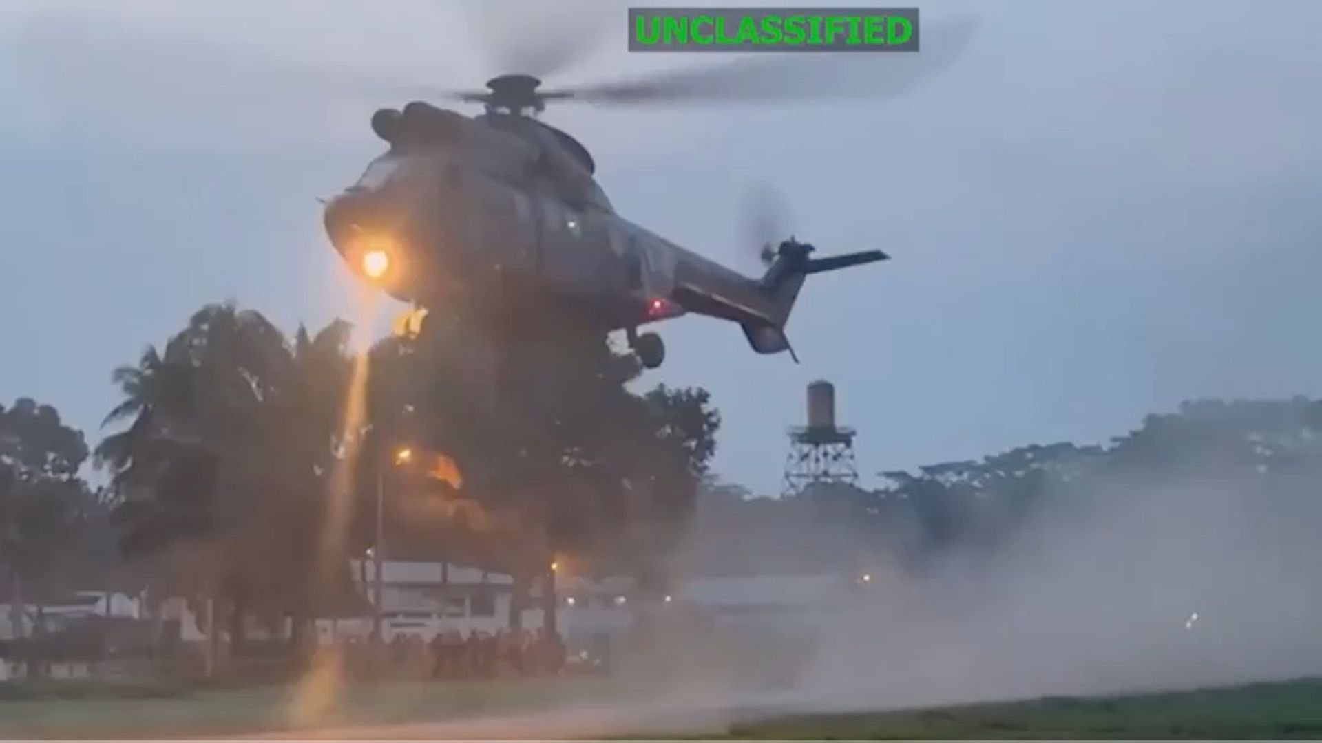 Military helicopter hovering low with its spotlight on, kicking up dust near a tree-lined area and buildings at dusk, with a water tower visible in the background. Marked as 