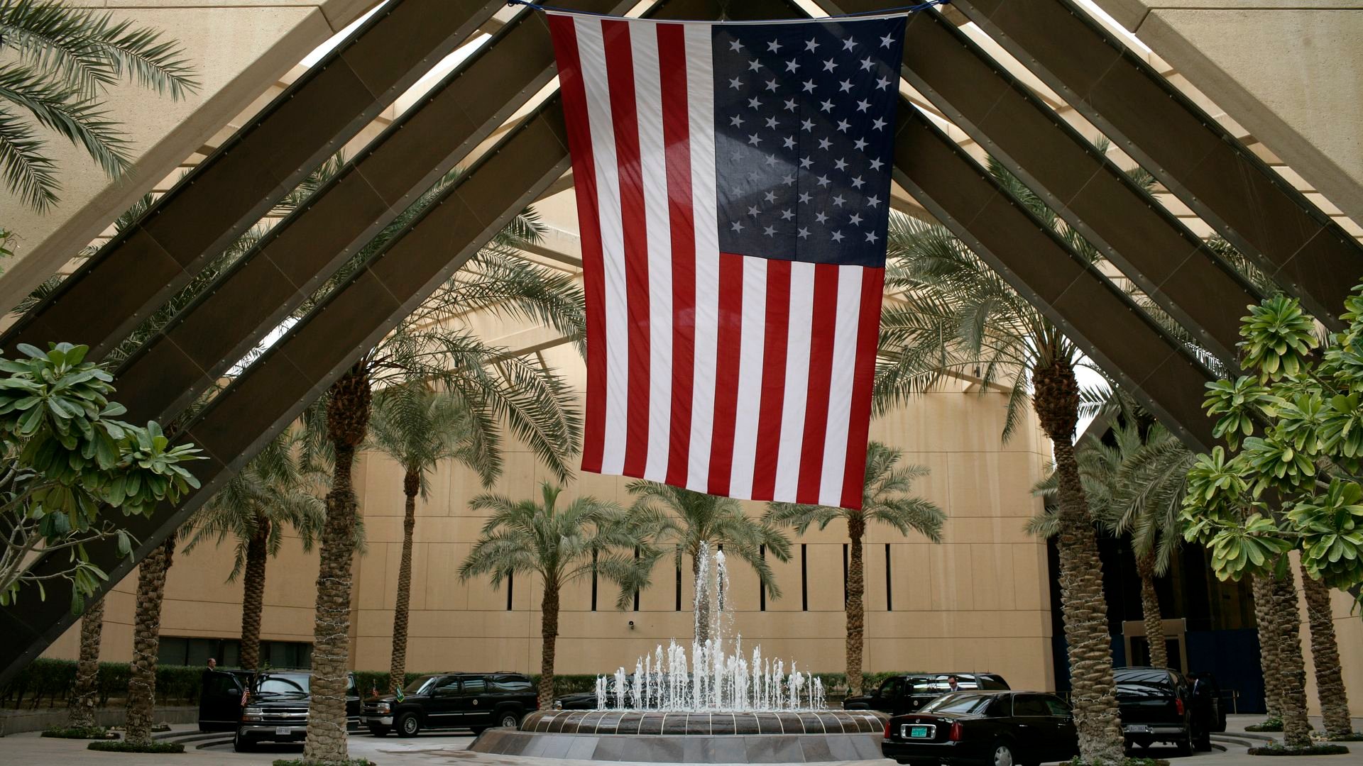 Large American flag hanging upside down inside a courtyard with palm trees, a water fountain, beige buildings, and several black vehicles parked around.
