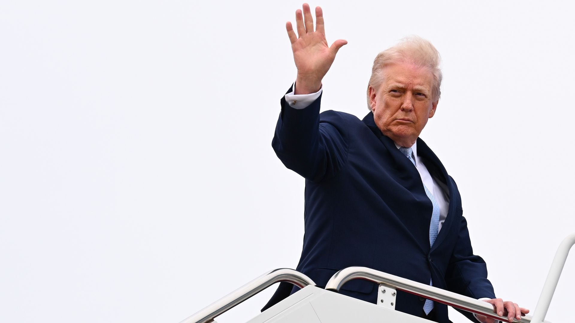 Donald Trump raises his hand while standing on the stairs of Air Force One against an overcast sky.