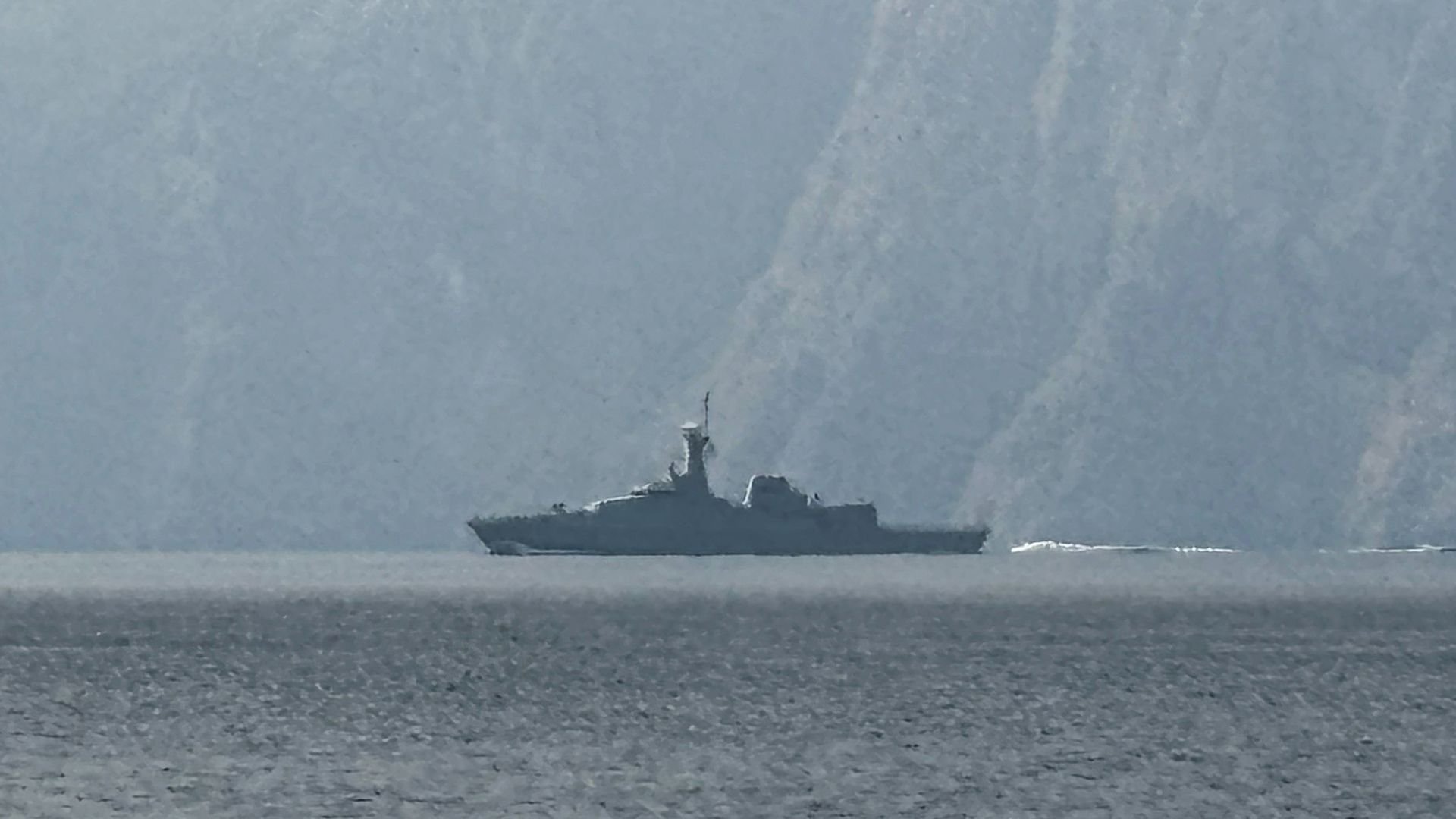 A naval vessel in a waterway with a mountain behind.