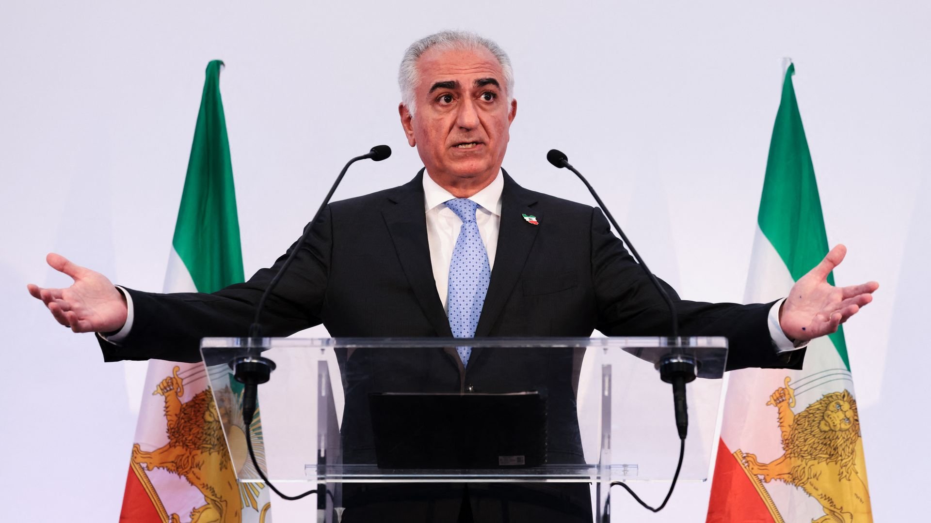 Pahlavi speaking at a podium with both hands raised slightly, wearing a black suit, white collared shirt and blue polka-dot tie