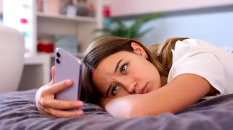 Getty Images A teenage girl lying on a bed, looking at her phone with a frustrated expression